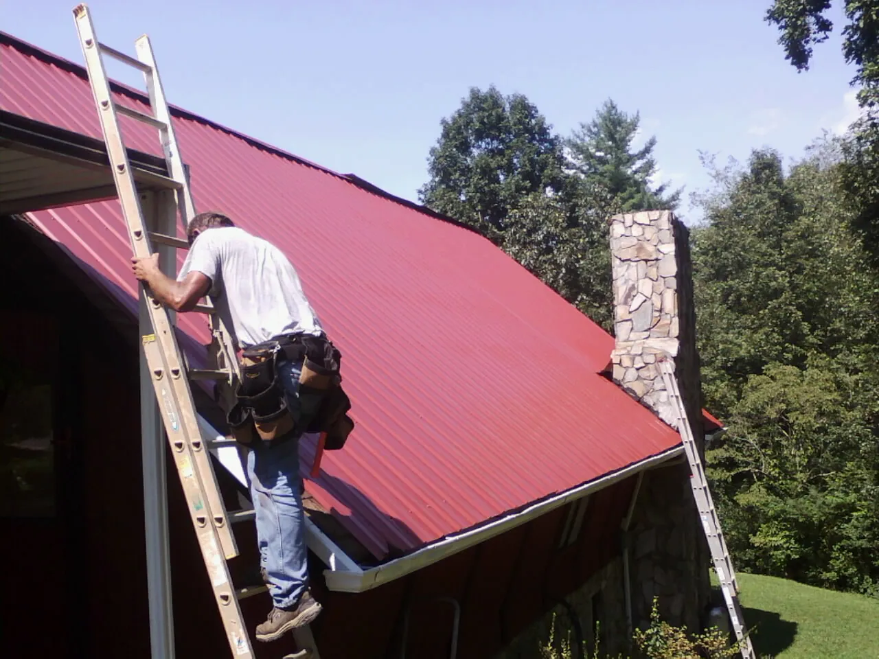 Roofer on a ladder beside a red metal roof and stone chimney — Soffit & Fascia Repair by Roofing Brothers in Seven Mile